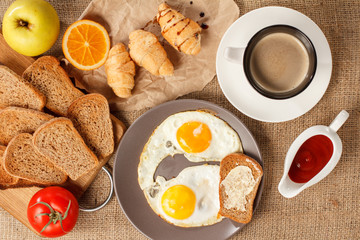 Plate with fried eggs and toast with butter, cup of coffee, croissants, orange, apple, bread on wooden cutting board, fresh tomato and ketchup in a sauce boat