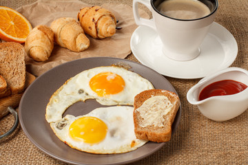 Plate with fried eggs and toast with butter, croissants, bread on wooden cutting board, fresh orange, cup of black coffee and ketchup in a sauce boat