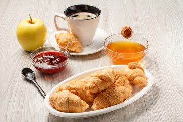 Fresh croissants on white porcelain dish, strawberry jam in bowl, apple, cup of black coffee with saucer, honey in bowl