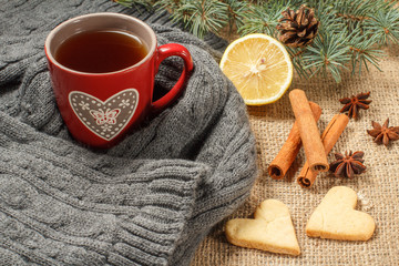 Obraz na płótnie Canvas Red cup of tea with pattern in heart shape wrapped up in wool scarf, slice of lemon, natural fir tree branches with cone, spices and gingerbread cookies