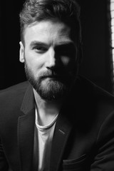Black and white close up portrait of young attractive man with dark hair, beard and mustache wearing dark jacket. Studio shot