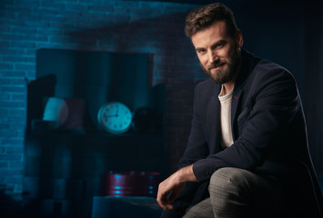 Studio portrait of handsome man with dark hair, beard and mustache wearing dark blue jacket, gray pants. Blue lighted brick background with clock and barrel