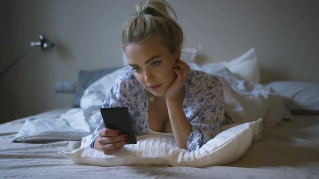 Lovely young woman with cute ponytail lying on comfortable bed and browsing smartphone in morning. 