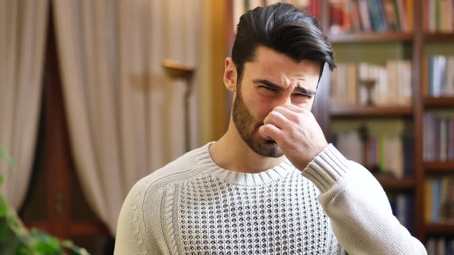 Portrait of caucasian young man closing nose with his hand because of bad smell. Looking at camera. At home, indoor