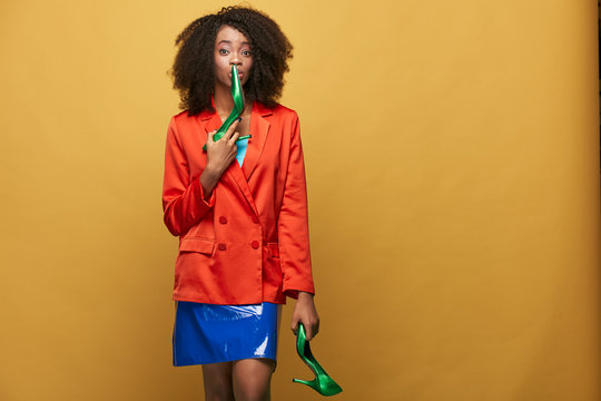 Funny colorful portrait of young african girl with afro hairstyle. Girl wearing  orange jacket and violet latex skirt holds green heels in her hand and kisses one of them. Studio shot.