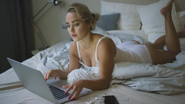 Pretty young woman in white panties and tank top lying on comfortable bed and using laptop in morning.