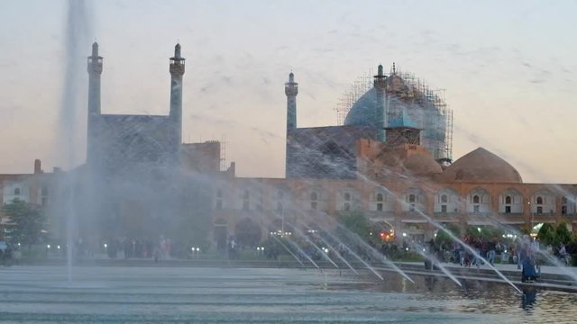 The walk along the evening Naqsh-e Jahan Square with scenic fountains and Shah Mosque (Royal or Imam Mosque) on the background, Isfahan, Iran