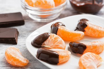 Slices of tangerine in chocolate served on a white plate, white wooden textured background.