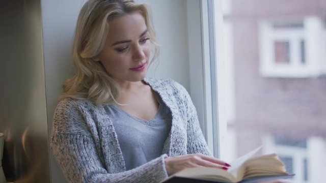 Pretty young woman in knitted jacket sitting on window sill and enjoying interesting book. 