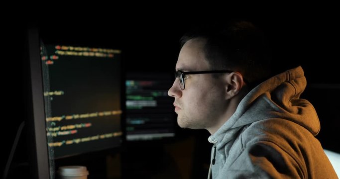 Male hacker in a hood works on a computer with maps and data on display screens in a dark office room