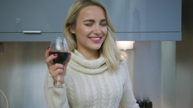 Lovely woman with blonde hair drinking red wine and cheerfully smiling while standing in kitchen. 