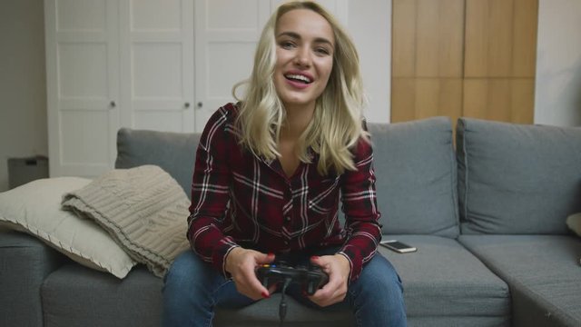 Excited young woman in checkered shirt holding controller and playing video game while sitting on comfortable sofa.