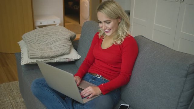Pretty young woman in casual outfit browsing laptop and cheerfully smiling while sitting on comfortable couch. 