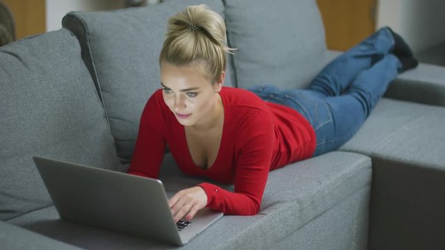 Gorgeous young woman in casual outfit lying on comfortable sofa and tying on laptop in living room. 