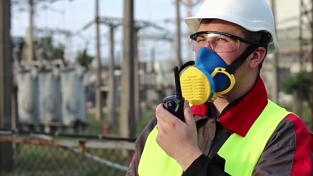 Electrician in respirator with portable radio transmitter on electric power station. Power engineering specialist in gas mask, goggles and hard hat on heat power plant communicates via radio station