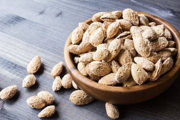 Salted almonds in a peel in a wooden bowl on the background of a dark tree. Selective focus.