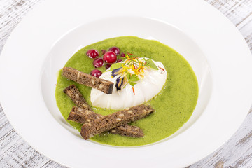 Poached egg, green healthy soup and slice of black bread with grains, close up in white plate