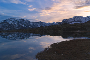 Fototapeta na wymiar High altitude alpine lake in idyllic landscape. Reflection of snowcapped mountain range and scenic colorful sky at sunset. Wide angle shot taken on the Italian Alps.