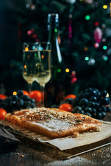 a festive New Year's table, cheese cake with chocolate, tangerines, grapes, biscuits, champagne and two glasses against the backdrop of a decorated Christmas tree with lights