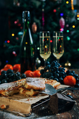 Fototapeta na wymiar a festive New Year's table, cheese cake with chocolate, tangerines, grapes, biscuits, champagne and two glasses against the backdrop of a decorated Christmas tree with lights