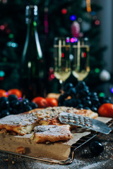 a festive New Year's table, cheese cake with chocolate, tangerines, grapes, biscuits, champagne and two glasses against the backdrop of a decorated Christmas tree with lights