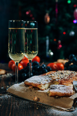 a festive New Year's table, cheese cake with chocolate, tangerines, grapes, biscuits, champagne and two glasses against the backdrop of a decorated Christmas tree with lights
