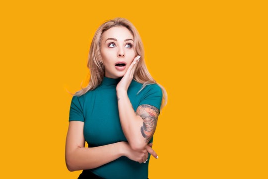 Surprised blonde woman with her hand on the face looking at the left part of camera. Yellow background in studio photo