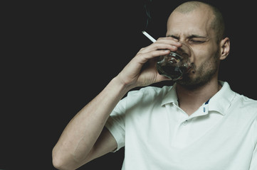 Bald man in white shirt drinking alcohol drink from the glass and smoking cigarette with black background not isolated