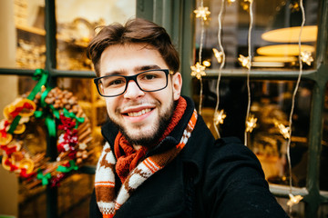 Handsome positive beard man blogger in glasses taking selfie outdoor near shop window with Christmas decorations of cozy shop in european city. Travel, tourism, holiday concept.