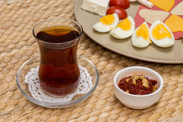 Close up of traditional Turkish tea and appetizer with blurred view of breakfast with cheese, salami, boiled egg, and tomato.