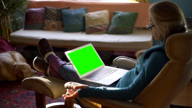 Steadicam motion side shot of pretty mature woman on green screen laptop computer with her feet up and reading out loud.