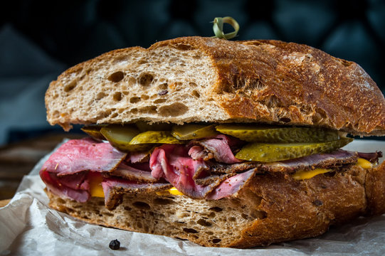 Pastrami sandwich on rye bread with pickles and mustard sauce, served on  wooden plate