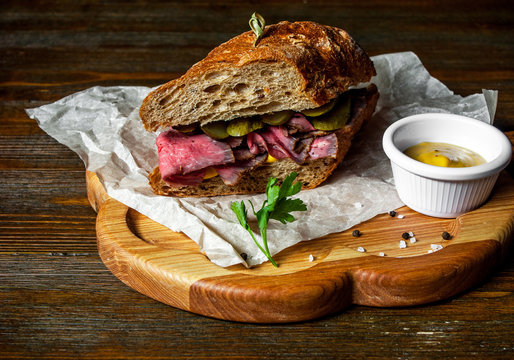 Pastrami sandwich on rye bread with pickles and mustard sauce, served on  wooden plate
