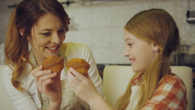 Portrait shot of the pretty and happy mother and daughter having fun while eating muffins at the kitchen table. Inside