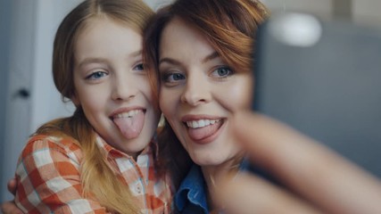 Portrait shot of the beautiful woman with young daughter making selfies on the smart phone, showing their tongues, hugging and laughing. Close up. Inside - Powered by Adobe