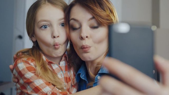 Close up of the smiling mother hugging young daughter and making funny selfies on the smartphone while grimacing. Portrait. Inside