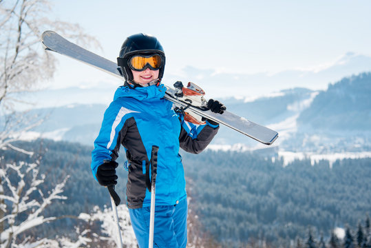 Portrait of happy female skier smiling to the camera, wearing skiing equipment, holding skis on the shoulder at winter ski resort copyspace happiness positivity vacation travelling