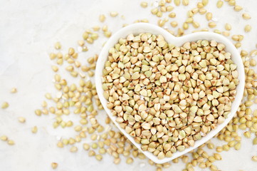 Heart shaped bowl of green buckwheat on buckwheat groat background, gluten free ancient grain for healthy diet, selective focus