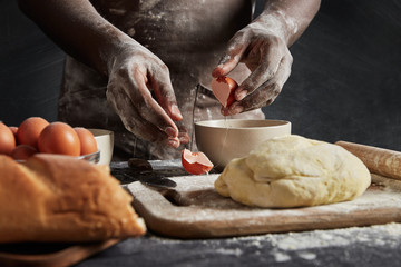 Cropped image of back man in apron, breaks eggs in bowl as prepares filling in buns, surrounded with prepared dough, rolling pin and white loaf, isolated over black background. Handmade concept