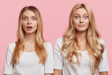 Horizontal shot of stupefied shocked two sisters stand close to each other, shocked to be hired for desired position in prosperous company, isolated over pink background. Two female companions