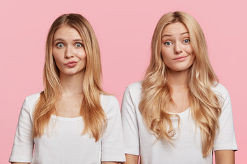 Puzzled blonde two females stand close to each other, curve lips, being uncertain about making decision, isolated over pink background. Beautiful sisters dressed casually don`t know what to do
