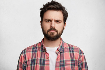 Serious gloomy man with thick dark beard, mustache and stylish hairdo, dressed in checkered shirt, being displeased as hears bad results of his work, isolated over white concrete background.