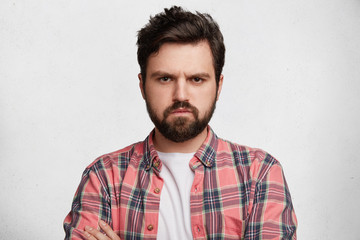 Handsome fashionable male has thick beard, dressed in checkered shirt, looks with grumpy expression at camera, being displeased after quarrel with girlfriend, isolated over white concrete wall