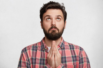 Fototapeta premium Serious bearded male prayer with palms pressed together, looks upwards, dressed casually, prays for good luck or health of relatives, isolated over white background. Religious guy worships success