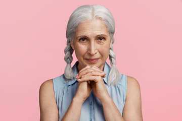 Glad beautiful senior woman looks happily at camera, keeps hands together, being pleased to hear positive news, isolated over pink studio background. Smiling delightful female with grey hair