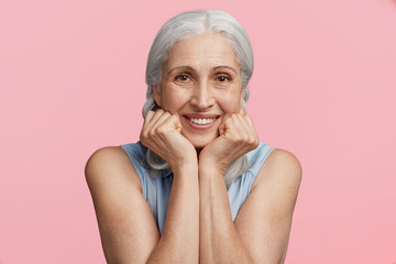 Smiling cheerful positive grey haired female with happy expression keeps hands under chin, looks...