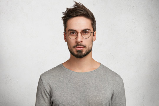 Stylish bearded male student wears round spectacles, has trendy hairstyle, looks confidently, thinks about coming session, isolated over white concrete background. People and human expressions
