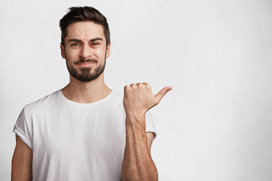 Portrait of displeased bearded young male model wears white t shirt, indicates with thumb aside, sees something unpleasant, isolated over studio background with copy space for your advertisment