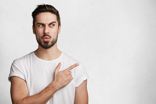 Puzzled hesitant bearded man with trendy hairdo, notices something awful, indicates with fore finger at blank copy space for your advertising content. Emotional fashionable guy in white studio
