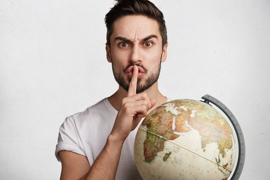 Annoyed young male teacher of geography holds globe, shows silence sign as demands calmness from pupils, isolated over white concrete wall. Bearded male traveler asks keep in secret new destination